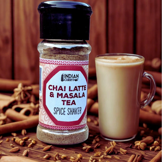 A bottle of Indian Curry Pot Chai Latte & Masala Tea Spice Shaker stands next to a glass mug filled with iced chai latte, surrounded by cinnamon sticks, whole spices, and star anise on a rustic wooden surface.