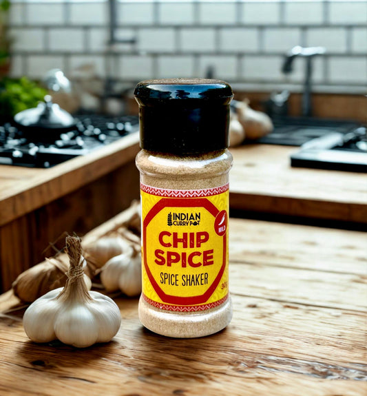 A shaker bottle of Indian Spice Farm Chip Spice stands on a wooden countertop in a kitchen setting, with garlic bulbs nearby and a blurred kitchen background.