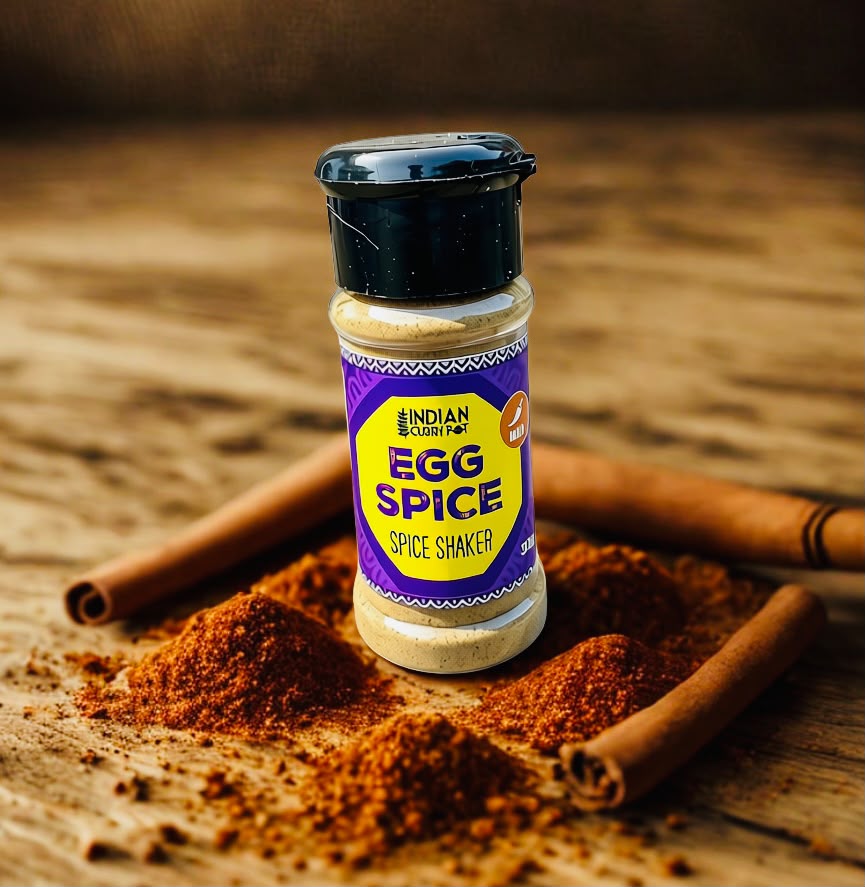 A bottle of Indian Spiced Egg Spice Shaker stands on a rustic wooden surface surrounded by cinnamon sticks and spilled brown spice powder. The label reads 'INDIAN SPICED EGG SPICE' and 'SPICE SHAKER'.