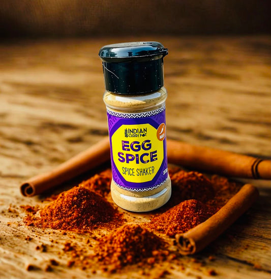 A bottle of Indian Spiced Egg Spice Shaker stands on a rustic wooden surface surrounded by cinnamon sticks and spilled brown spice powder. The label reads 'INDIAN SPICED EGG SPICE' and 'SPICE SHAKER'.
