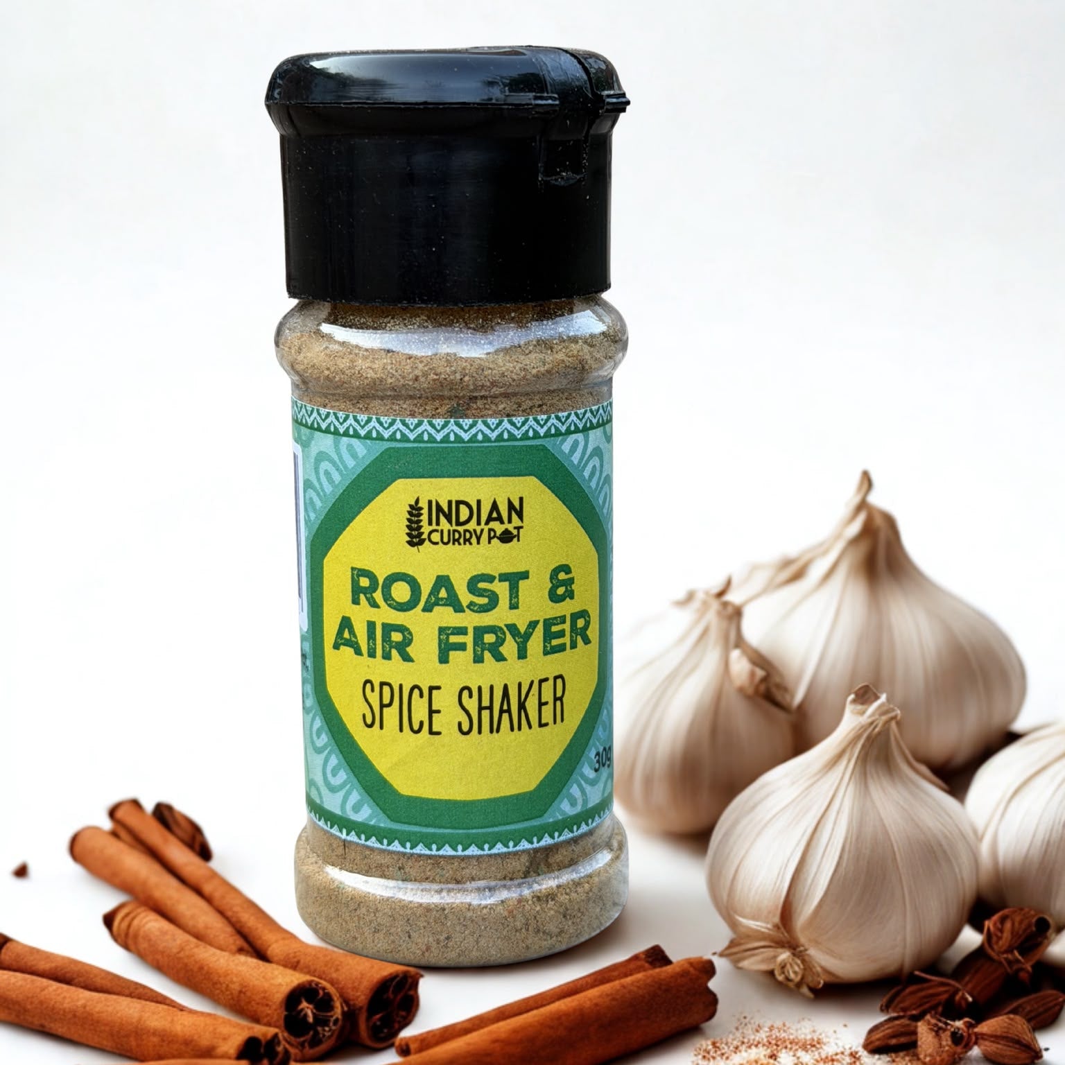 A bottle of Indian Curry Roast & Air Fryer Spice Shaker stands on a white surface surrounded by cinnamon sticks, star anise, and garlic bulbs, with a light gray background.