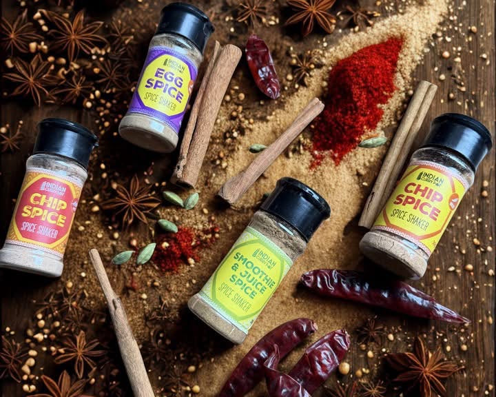Four spice shaker bottles arranged on a rustic wooden surface with cinnamon sticks, star anise, dried red chilies, and a spoonful of red chili powder. A label at the top reads 'spice shakers'.